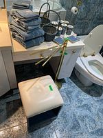Vanity stool on casters with white cushion, stacked gray hand towels, wicker basket, gold-toned adjustable floor lamp stand, and part of toilet in bathroom setting.
