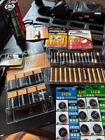 Photo of various batteries laid out on table including Duracell 9V, button cells in packages, and flashlights.