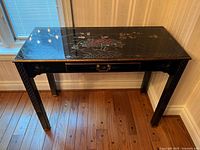 Front view of the black lacquered table showing floral and bird hand-painted detail on top along with drawer and leg details.