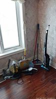 Wide shot showing multiple cleaning items near a window including brooms, handheld vacuum, and mop