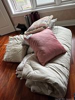Comforter set with accent pillows stacked on wooden floor near window