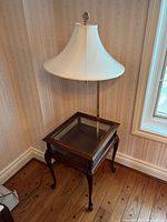 Full view of display table with attached lamp in a room corner showing table wood finish, glass top, and white shade lamp.