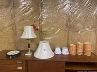 Clock, lamps, and lampshades displayed on a wooden surface with plastic-covered wall background.