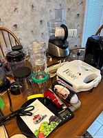 Photo shows digital kitchen scale with black glass surface, Magic Bullet jars and motor blades, sandwich maker, and other small appliances on wooden table.