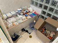 Assorted sealed medical supplies arranged on bathroom ledge and floor including gauzes, medication organizers, sealed boxes, plastic wash basin, and first aid dressings.