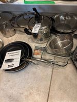 Photo showing stainless steel Langostina pressure cooker, three Henckels black granite pans, glassware, metal pots and pans with lids, metal racks, and a wooden cutting board.