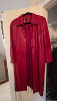 Full view of red leather trench coat hanging on door, shows length, collar, buttons, and overall condition