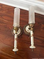 Two matching wall-mounted light fixtures laying on wooden floor showing ceramic bases with floral decal, metal hardware, round wooden backplates with floral pattern, and frosted glass shades.