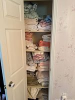 View of linen closet shelves showing multiple stacks of folded towels, sheets, and blankets in neutral and pastel colors.