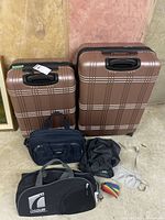 Two matching bronze hard sided suitcases with white stripes, one smaller than the other, placed against a basement wall, along with several additional travel bags and accessories.