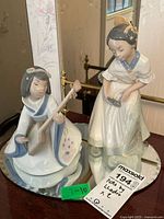 Two porcelain Nao by Lladro figurines on a mirrored brass base, one seated playing an instrument and the other standing holding a mirror, showing overall lot with tags.