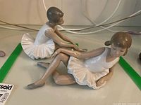 Two Nao by Lladro ballerina figurines sitting with legs posed on a glass surface. Figurines have white tutus, gray ballet shoes, and brown hair in pigtails.