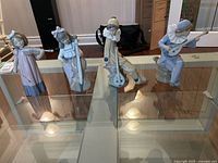 Four porcelain musician figurines on a glass shelf, showing all pieces together with reflective surfaces and background boxes.