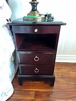 Front view of one wood nightstand showing small upper drawer with golden knob, open ledge, two lower drawers with ring pull handles on wooden floor