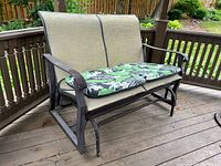 Outdoor gliding loveseat showing side angle with two floral cushions on seat and beige seating mesh with some wear.