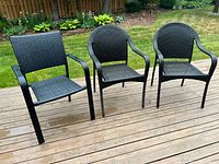 Front view of three black patio chairs placed on wooden deck next to grass lawn. One chair with square back on left, two chairs with rounded backs on right.