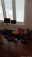 Wide shot showing multiple purses and wallets including black handbags, yellow handbag, pink handbag, patchwork purse and wallets placed on the floor by the window with blinds.