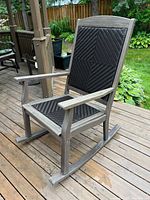 Side angle showing the wooden frame with grey finish and the woven black seat and backrest of the rocking chair on a wooden deck.
