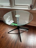 Round glass top side table with black metal tripod base, showing top surface and slight scratches on glass.