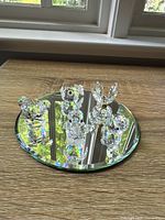 Five Swarovski crystal figurines displayed on a large round beveled mirror reflecting light and colors, showing fish, butterfly, and two ducklings.