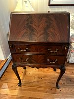 Front view of dark wood vintage secretary desk showing slant front, two drawers with brass handles, cabriole legs ending in pad feet, wood grain pattern and patina.