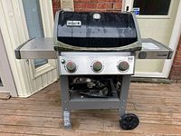 Front view of the Weber Spirit II barbecue grill showing the black lid with temperature gauge, three control knobs, and attached side shelves.