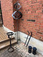 Photo showing kinetic wind spinner, tall plant holder, smaller plant holder, two solar lanterns, and metal sundial against brick wall.
