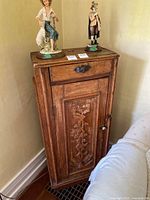 Full view of antique wooden cabinet with drawer and door with carvings placed against the wall with figurines on top.