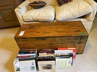 Front view of the wooden box placed on a carpeted floor next to a white couch, showing the rectangular shape and natural wood grain with some dark patches.