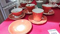 Set of 6 coral/red Aynsley teacups with matching saucers and one additional saucer on a red tablecloth.