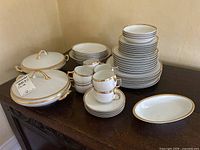 Full display of the china set on a dark wooden sideboard showing stacked plates, bowls, cups, saucers, and two covered serving dishes with gold handles.