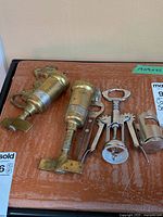 Photo showing two brass vintage style spring-loaded corkscrews, a wing corkscrew and part of another corkscrew on a brown surface.