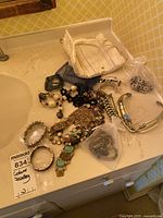 Overview of costume jewelry pieces including necklaces, multiple bracelets, and fabric storage bag placed on a bathroom counter.