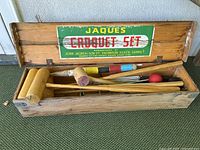 Wooden box open showing croquet mallets, balls and metal wickets inside with Jaques Croquet Set colorful label on the lid.