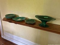 Overview of nine green dishes on wooden shelf: stack of small plates, serving platter, shallow bowl, pedestal compote