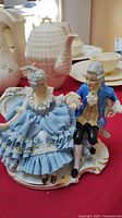 Porcelain figurine of 18th-century couple with man in blue coat and woman in blue ruffled dress, showing fabric lace and floral details