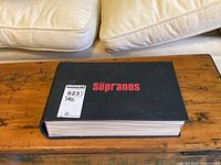 Closed hardcover style DVD box set with black cover and red title text 'The Sopranos'.