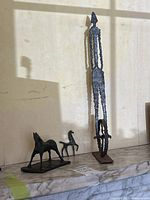 Wide shot of three metal figurines placed on marble surface against a white wall. Shows tall cyclist and two horse sculptures.