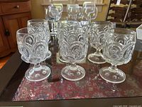 Six clear glass goblets arranged on a reflective glass surface showing intricate embossed circular and floral patterns on the bowls and star-like patterns on the stems' base.