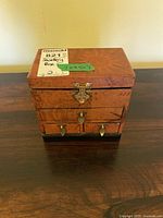 Front view of wood jewelry box showing natural wood finish, three drawers each with gold knobs, and decorative clasp on front top.