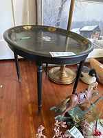 Oval metal top table with black finish and gold floral painted decoration around the edge and center. Visible scratches and paint wear present on the metal surface. Wood legs are turned and finished dark.