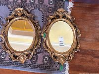 Two paired oval mirrors with ornate antique carved wood frames resting on a patterned rug and wooden floor.