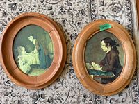 Two oval paintings laying on a patterned carpet. Both have wooden frames. One shows a woman serving a child; the other a girl reading.
