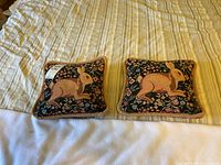 Two matching square decorative cushions placed side-by-side on a striped and white fabric background, showing size difference visually.