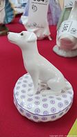 Front angled view of the white porcelain dog figurine seated on a patterned purple and white base.