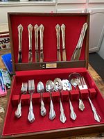 Open wooden box with red velvet lining showing full silverware set including knives in lid slots, and forks, spoons, serving utensils in bottom slots.