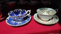 Photo of two teacups and saucers on red fabric: left is the Coalport blue floral pattern, right is Paragon with gold leaf design.