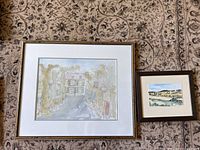 Photo shows both watercolours laying on a patterned rug, one large and framed in gold with white matting depicting a building and trees, the smaller scenic painting framed in dark wood