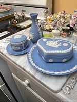Photo showing two Wedgwood blue jasperware trinket boxes with white relief decoration, a matching Jasperware vase, and two plates, all arranged on a countertop.