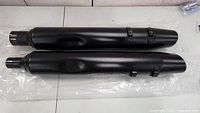 Top view of both Harley Davidson mufflers laid side by side, showing length, mounting brackets and black finish.